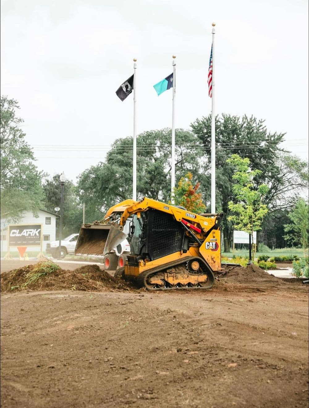 Caterpillar skid steer loader clearing dirt near flagpoles and landscaping.