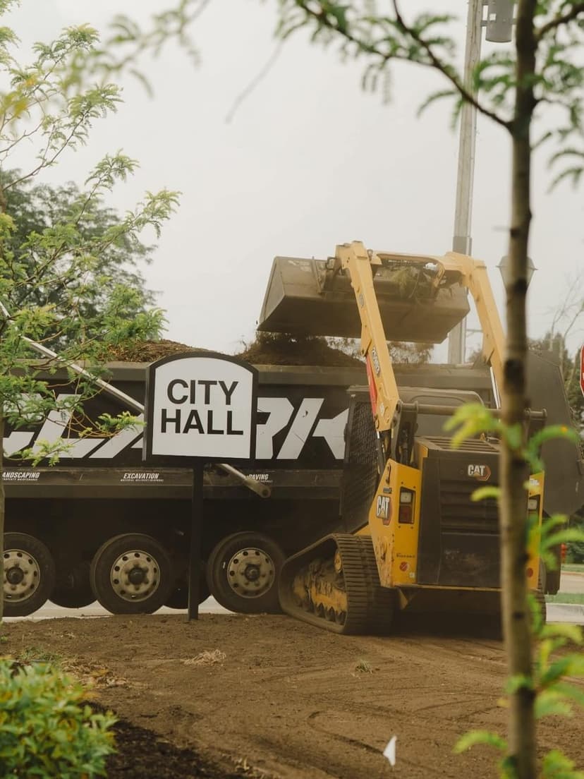 Excavator loading soil into a dump truck near a City Hall sign, surrounded by greenery.